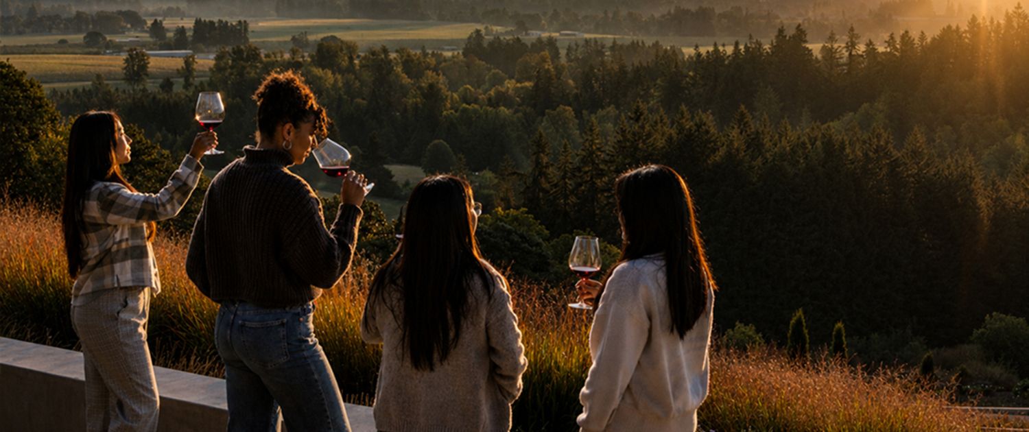 Four women stand outdoors at sunset, holding glasses of wine and looking out over a scenic landscape of trees, fields, and distant hills.
