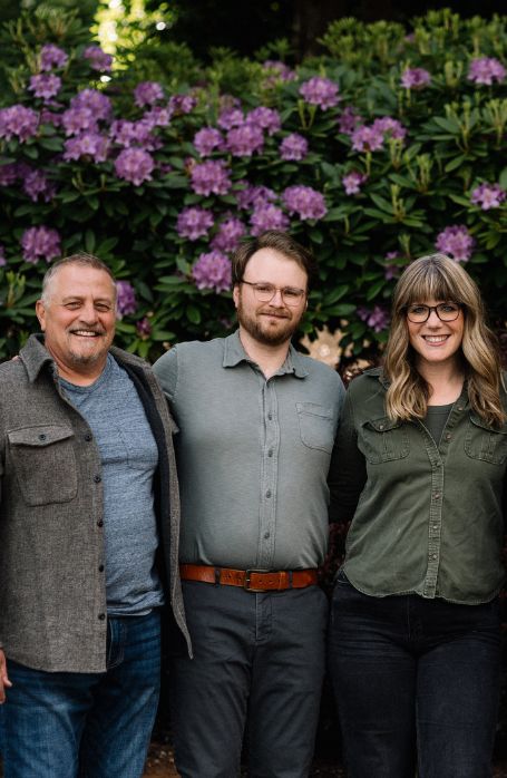 Three people stand close together, smiling in front of a lush green bush with blooming purple flowers. They are dressed casually and appear relaxed outdoors.