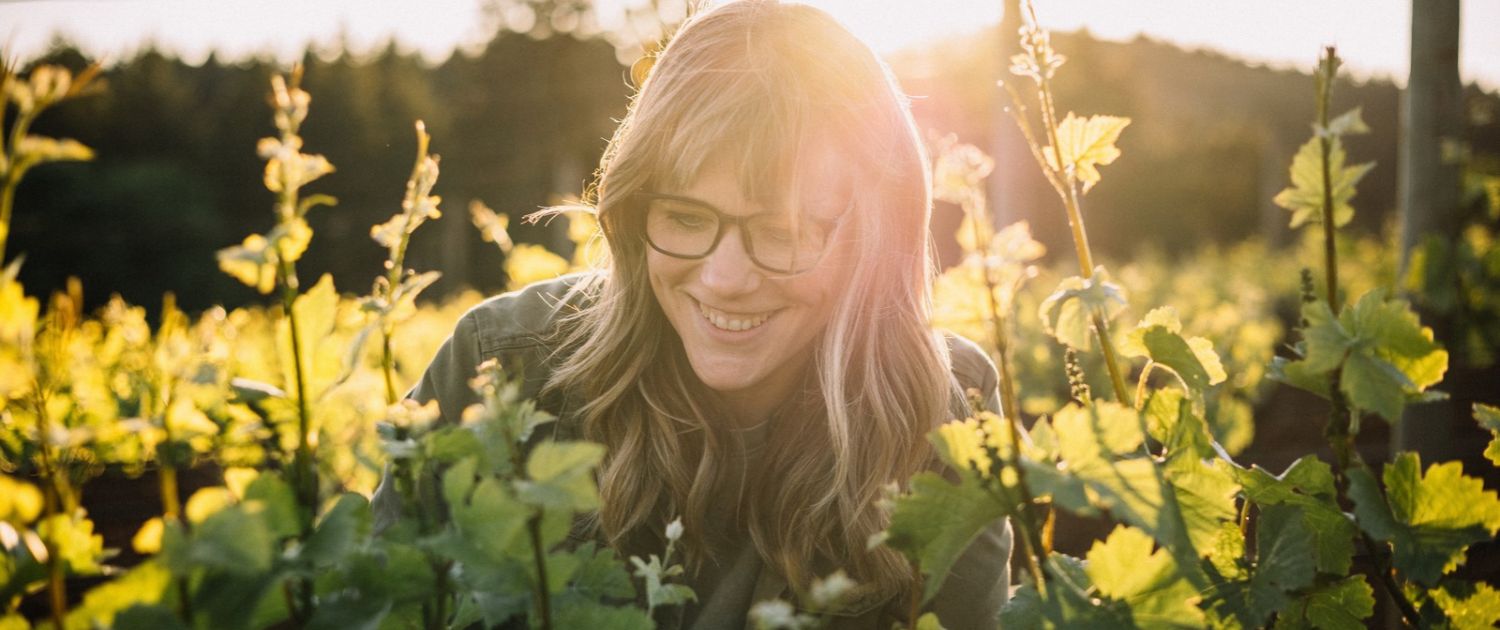 A woman wearing glasses smiles as she tends to grapevines in a vineyard at sunset.