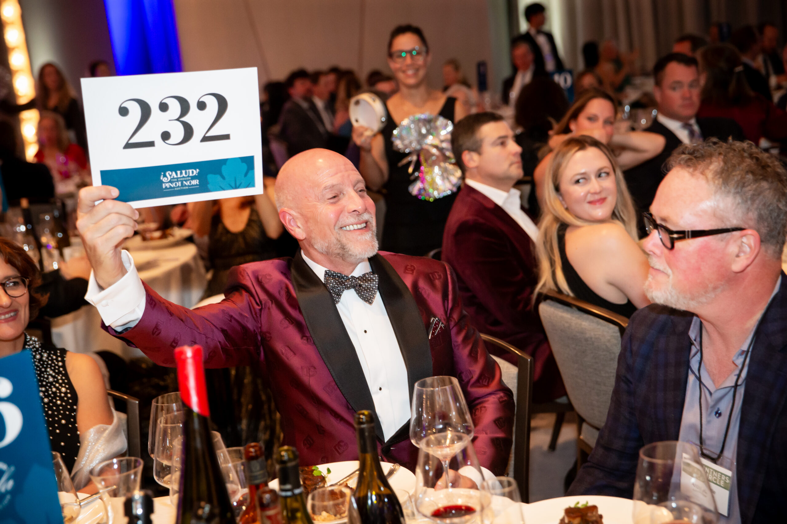 A smiling man in a red suit holds up a card with 232 at a formal event. People in elegant attire sit around tables with wine glasses and bottles, appearing engaged and cheerful.